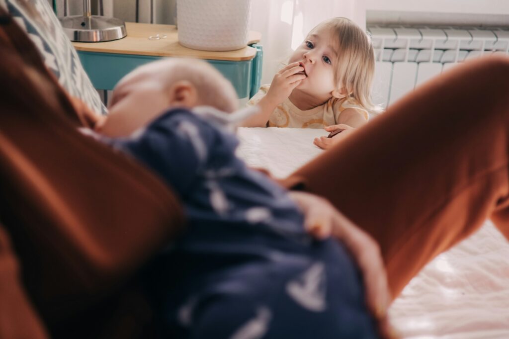 toddler girl looking at her mother breast-feeding an infant baby