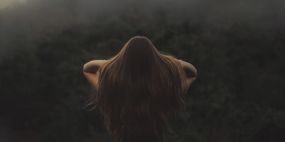 La Lune influence-t-elle vraiment la pousse des cheveux ? Back view of a woman with long hair, standing in a misty forest.