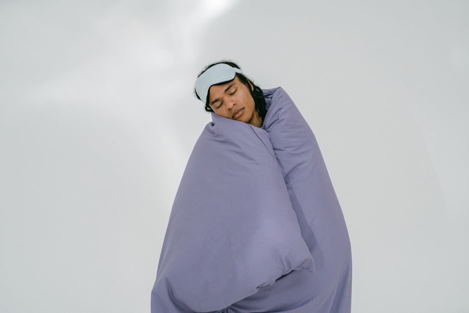 Les rêves lucides sont-ils scientifiquement prouvés? A person peacefully sleeping with an eye mask, wrapped in a purple blanket on a white background.