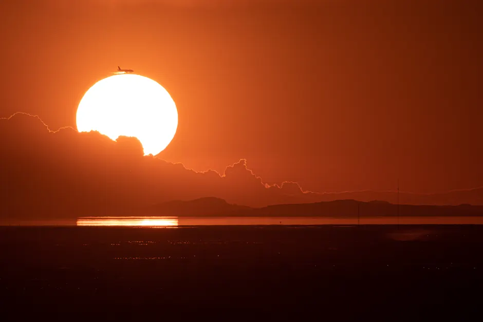 Le Soleil finira-t-il vraiment par exploser? Silhouette of airplane passing giant sun at sunset over the ocean, creating dramatic sky and reflections.