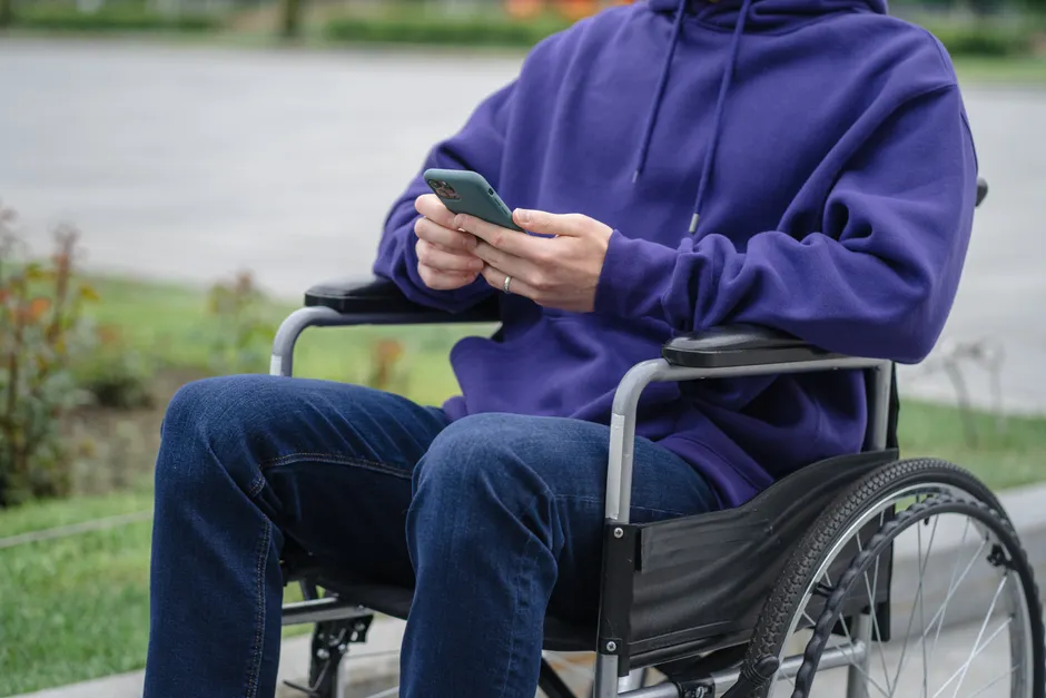 A person in a wheelchair texting on a smartphone while wearing a purple hoodie, outdoors.