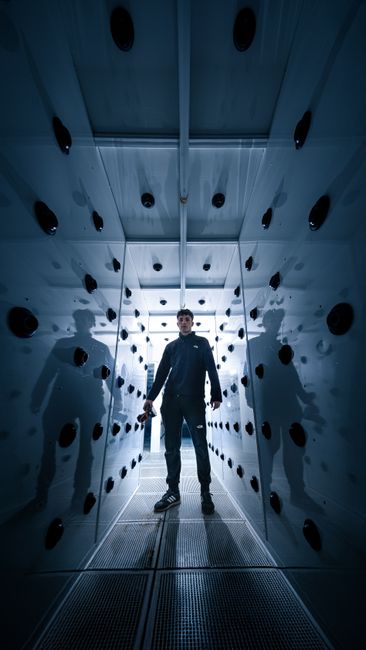 A person stands in a futuristic, reflective industrial chamber with blue lighting, creating a sci-fi atmosphere.