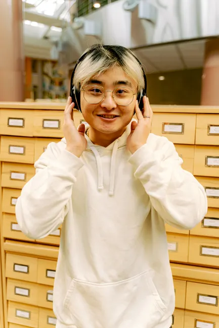 A smiling person with headphones in a white hoodie standing in front of card catalog drawers indoors.