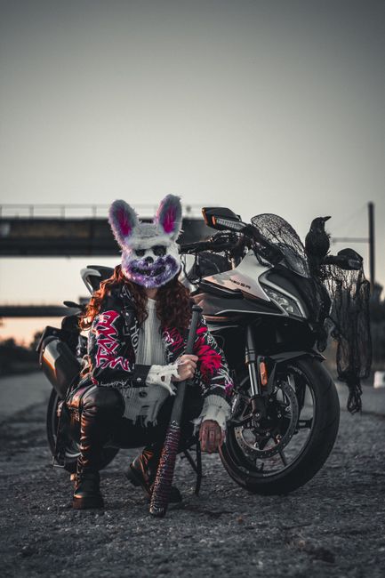 A striking image of a person in a bunny mask posing with a motorcycle outdoors, featuring a crow on the bike.