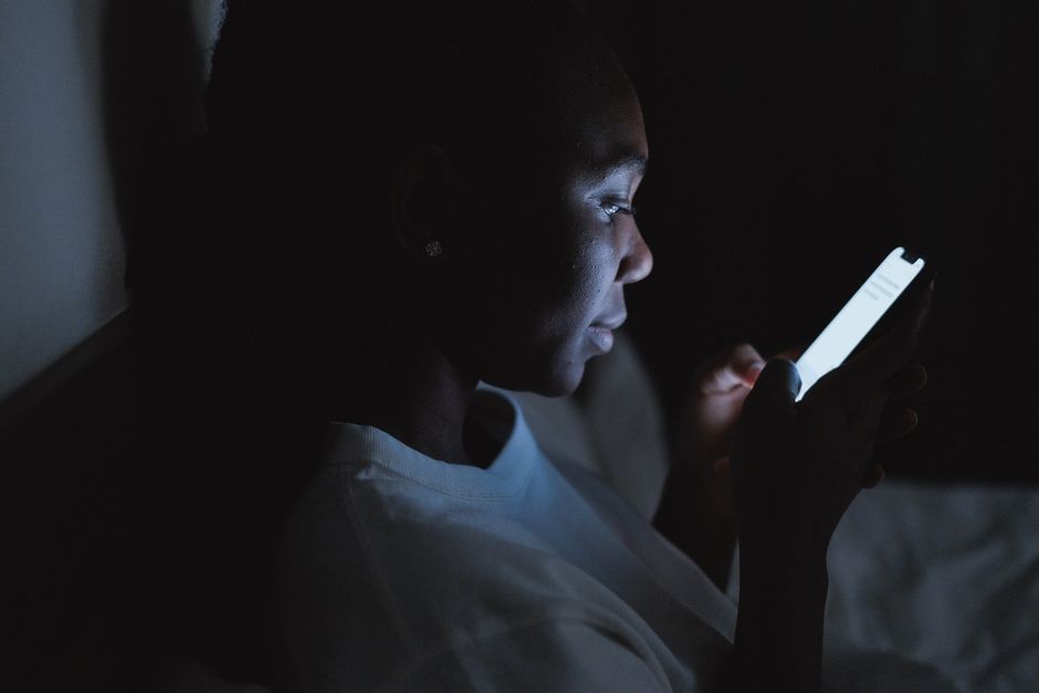 La lumière bleue empêche-t-elle vraiment de dormir? Un effet réel, souvent exagéré A woman using a smartphone in a dark bedroom, lit by the screen glow at night.