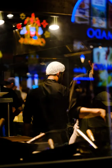 Back view of a chef in a bustling urban restaurant kitchen at night with neon reflections.