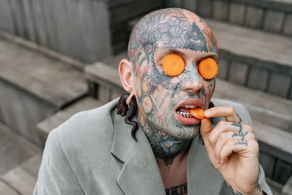 Les carottes améliorent-elles vraiment la vision nocturne? Bald tattooed man with bright tattoos and carrot slices on face and in hand, outdoors.