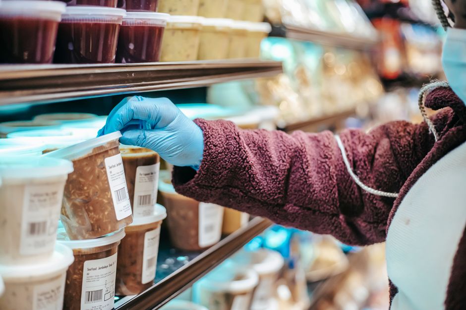 Peut-on vraiment manger un aliment après sa date de péremption? Crop anonymous female customer in protective mask reading label on frozen food in plastic container in grocery store
