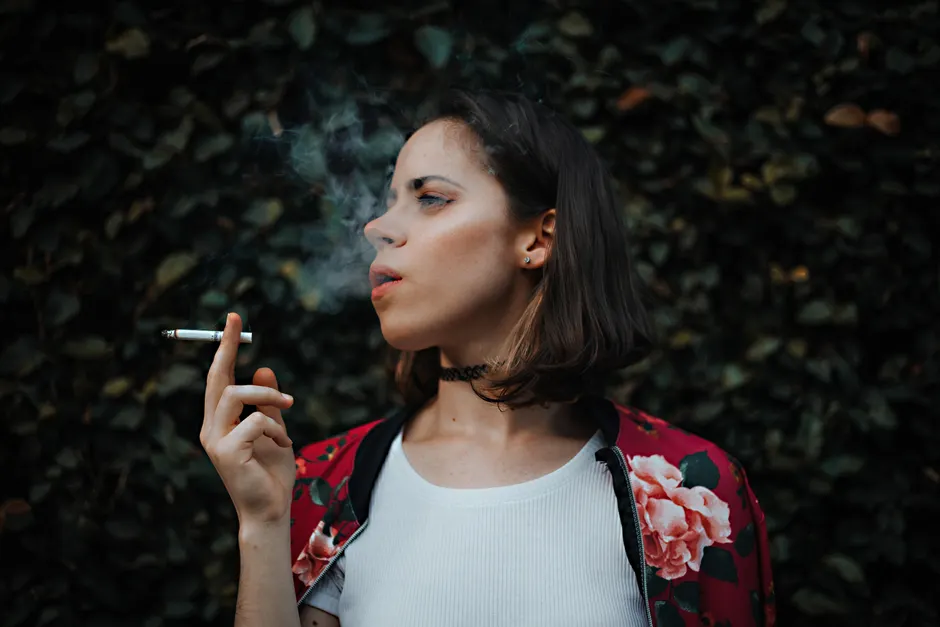 L’hypnose peut-elle vraiment aider à arrêter de fumer? Portrait of a woman smoking outdoors, clad in a floral jacket, exhaling smoke against a leafy backdrop.