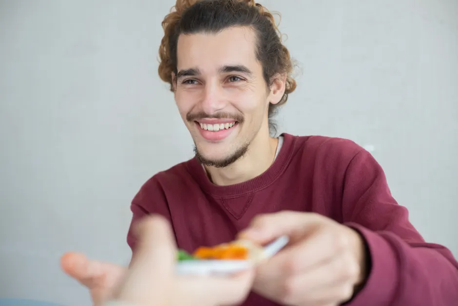Smiling young man with beard and mustache sharing a meal indoors, expressing warmth and friendliness.