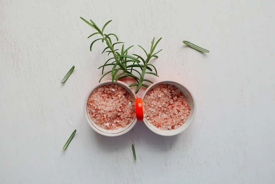Le sel rose de l’Himalaya est-il vraiment meilleur pour la santé? Top view of pink Himalayan salt and fresh rosemary in bowls on white table.