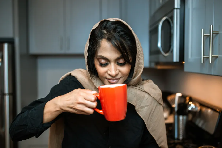 Les odeurs peuvent-elles vraiment réveiller des souvenirs oubliés? Woman in hijab delighting in the aroma of coffee from a red mug in a modern kitchen.