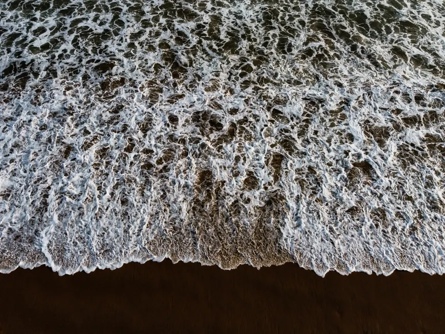 A captivating top-down view of ocean waves crashing onto the sandy shoreline, creating a textured sea foam pattern.