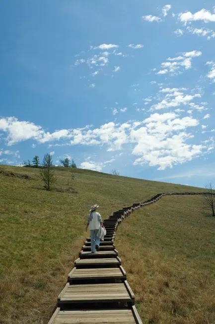 A person walks on a rustic wooden boardwalk through a grassy hillside under a bright blue sky.