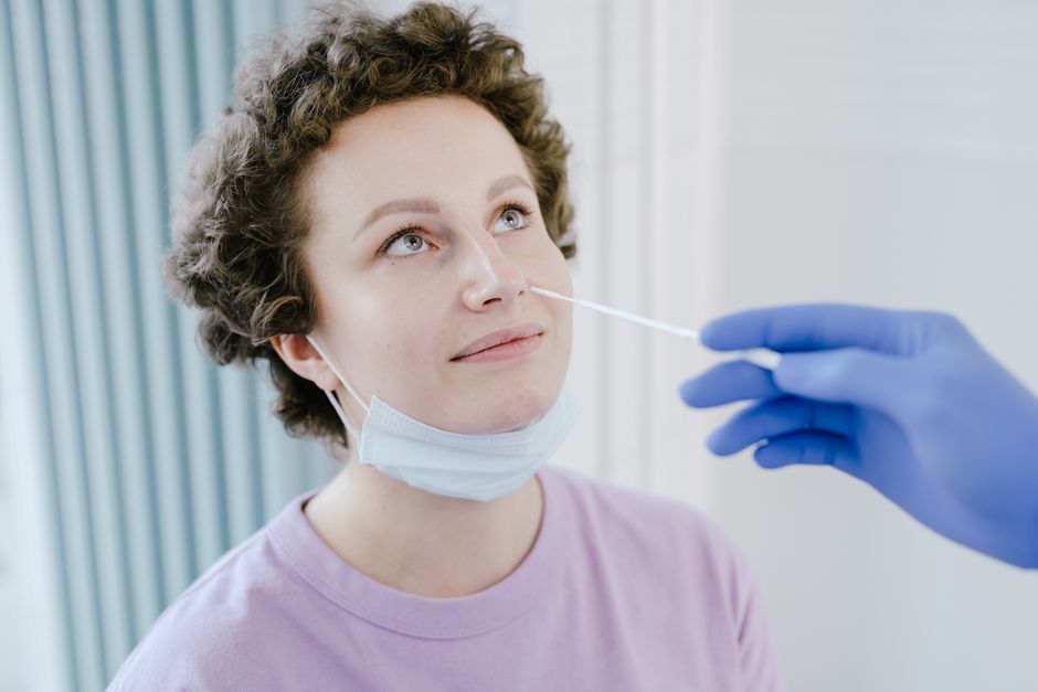 Peut-on vraiment attraper deux fois le même virus? A woman in a medical facility receiving a nasal swab COVID-19 test, wearing a mask, smiling.