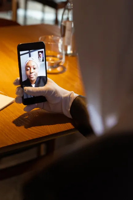 A woman with a headscarf has a video call on her smartphone while seated at a wooden table.