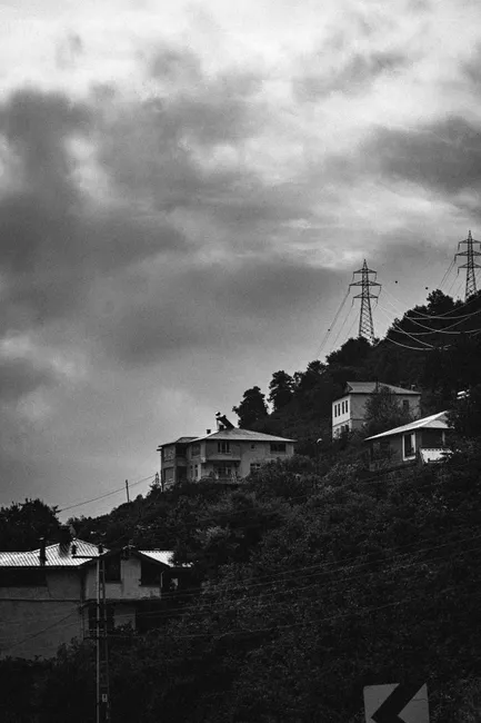Black and white image of hillside with houses under stormy clouds and power lines.