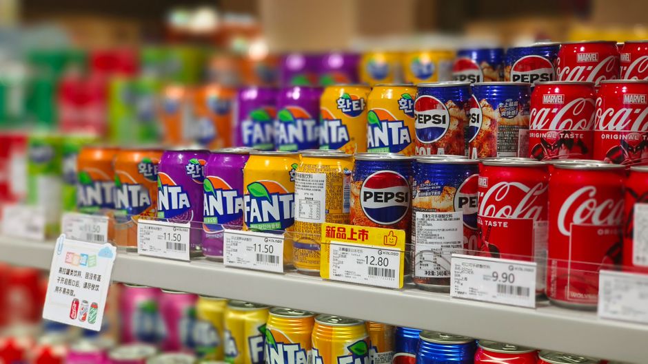Les supermarchés mettent-ils les produits chers à hauteur des yeux? Colorful soda cans including Fanta, Pepsi, and Coca-Cola arranged on supermarket shelves, price tags visible.