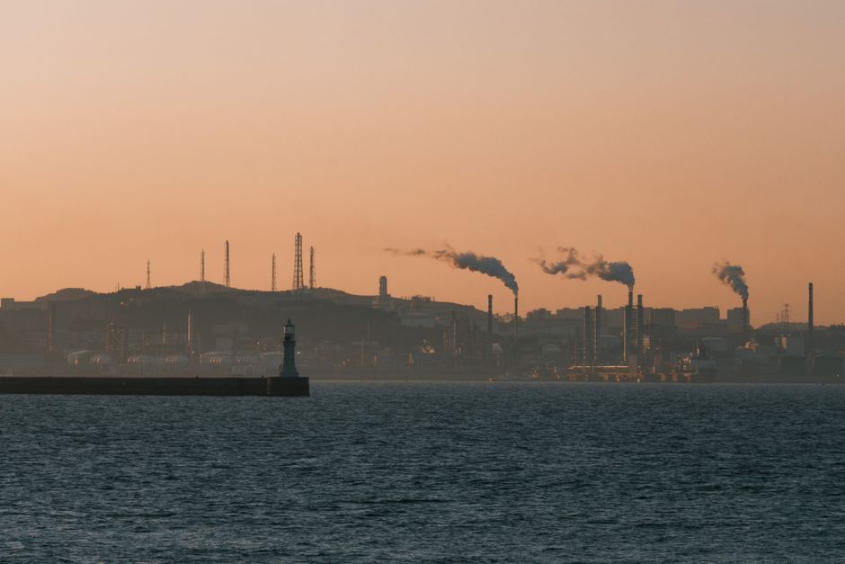 Factory skyline emitting smoke over the ocean at sunset, under an orange sky.