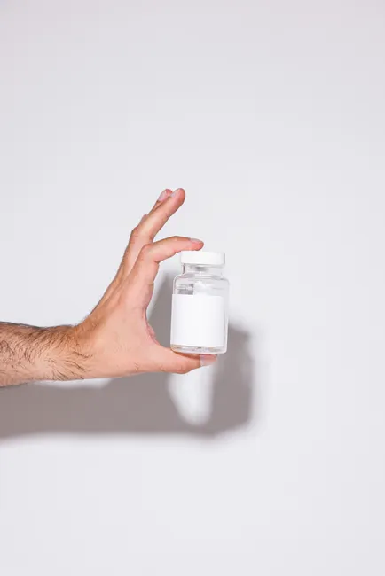 Hand holding a transparent bottle with white label against a plain background.