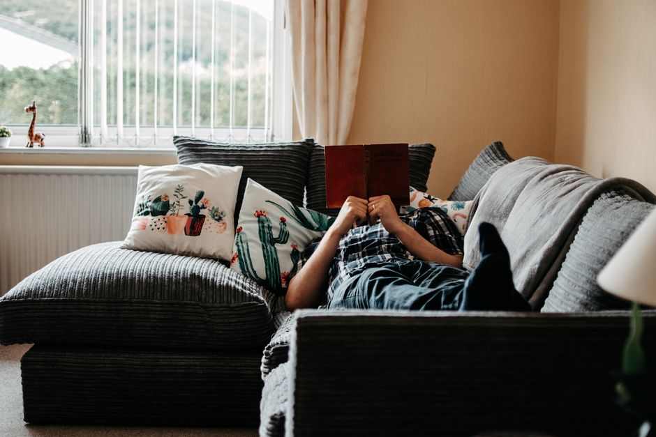 Person relaxing and reading on a cozy sofa at home, surrounded by cushions and natural light.