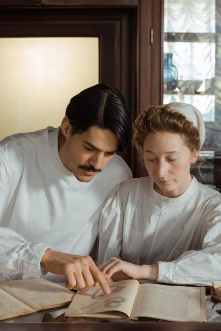 Two pharmacists in vintage attire examining medical books in a classic pharmacy setting.