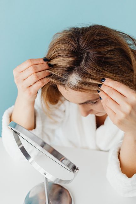 Woman in bathrobe checking grey hair roots in front of a mirror indoors.