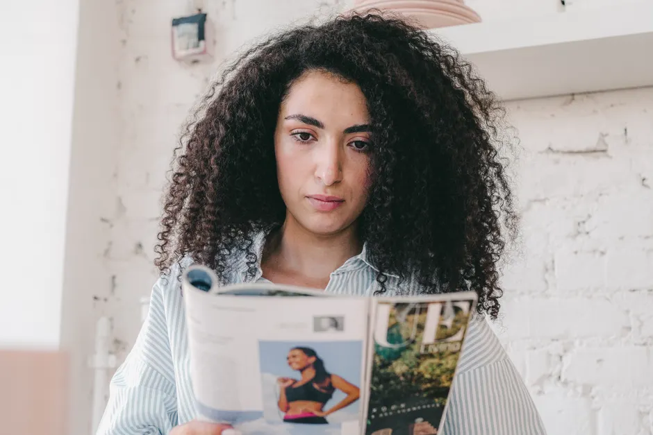 Young woman with curly hair reading a magazine indoors, focused and relaxed.